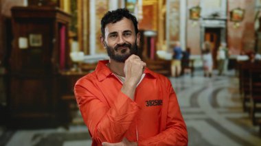 Turuncu tulumlu İspanyol sakallı bir adam Katolik kilisesinin içinde duruyor, içsel olarak poz veriyor, inanç, kefaret ve iç gözlem temaları öneriyor..