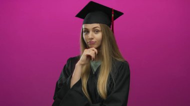 Mezuniyet şapkalı ve cüppeli bir kadın pembe arka planda yalnız düşünüyor.