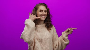 Bir telefon konuşması yaparken gülümseyen ve parlak pembe bir arkaplanı işaret eden, neşeli ve cana yakın bir ifadeyle rahat bir kazak giyen bir kadın..