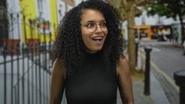 Gözlüklü genç bir kadın neşeli ifadesini ve kıvırcık saçlarını sergileyerek canlı şehir ortamında renkli binalarla dolu canlı bir sokakta gülümsüyor..