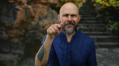 Mavi tişörtlü kel bir adam kayalık bir arka planı olan açık şehir ortamında ciddi bir şekilde el kol hareketi yapar. Yoğun ifadesini ve çekici tavırlarını yakalar..