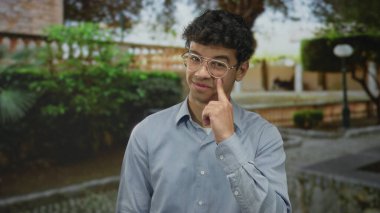 Gözlüklü ve mavi gömlekli genç İspanyol yeşil park ortamında parmak yanağa..