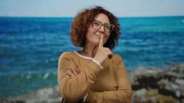 Orta yaşlı İspanyol bir kadın, deniz kenarında, mutlu bir ifadeyle, gözlük takıyor ve kahverengi bir süveter giyiyor, canlı mavi bir deniz ve gökyüzünün zeminine karşı..