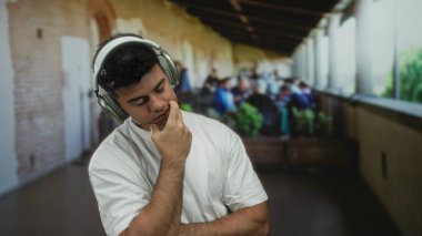 Kulaklık takan genç bir adam, arka planda bulanık insanlarla açık bir restoranda terasta kafa yoruyor, canlı bir şehirde düşünceli bir an sergiliyor..