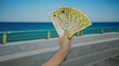 Arka planda açık mavi gökyüzü olan deniz kenarındaki gezinti alanının yanında Filipinli pesoları tutan adam plajda rahatlamış bir havayla para sergiliyor..