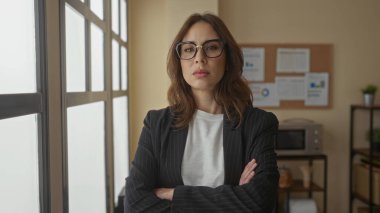 Gözlüklü kadın, büyük pencereli bir ofiste, spor ceket giymiş, profesyonel bir iş ortamını ön plana çıkarıyor..