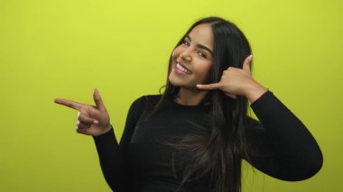 Genç kadın jestleri, neşeli bir şekilde gülümseyen parmaklarıyla beni arar. Canlı sarı bir arkaplan, iletişim ve mutluluğu iletir..