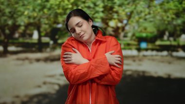 Turuncu hapishane tulumlu kadın yeşil ağaçlarla çevrili bir açık hava parkında kendini kucaklıyor, içsel gözlem ve sükunet duygularını aktarıyor..