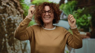 Parkta neşeyle dans eden bir kadın kıvırcık saçlı, parlak bir enerji sergiliyor, gözlük takıyor ve yemyeşil bir ortamda kahverengi bir süveter giyiyor..