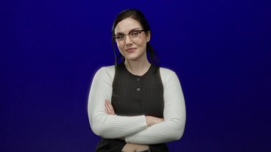 Gözlüklü bir kadın canlı mavi bir arkaplan karşısında güvenle gülümsüyor, esmer saçlı ve katlanmış kollu sıradan bir stil sergiliyor, coşkulu bir güzelliği ve zarafeti var.