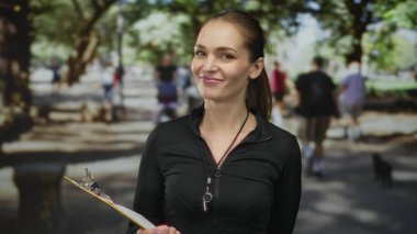 Beyaz bir kadın sarı bir pano tutuyor ve parktaki ağaçların oluşturduğu yol boyunca bir panoyu işaret ediyor; sağlık rehberliği güveni.