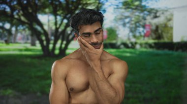 Çıplak göğsü olan bir adam sabah güneşi altında yeşil bir parkta eliyle omzuna hafifçe dokunur. Huzur..