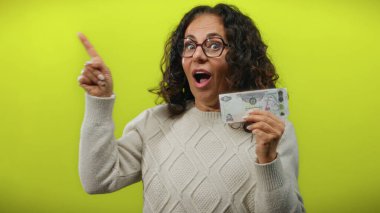 Uae 'den orta yaşlı bir kadın Dirham banknotunu tutarken izole edilmiş sarı arka plana şaşırmış görünüyor..