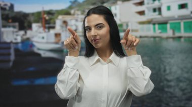 Beyaz tişörtlü bir kadın elleriyle kalp hareketi yapıyor ve canlı limanda öpüşüyor. Arka planda güneşli açık hava ortamının altında tekneler var..