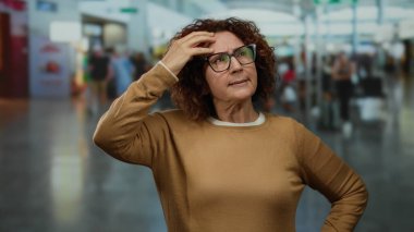 Havaalanı terminalinde hayal kırıklığına uğramış, orta yaşlı, kıvırcık esmer, gözlüklü, İspanyol görünümlü, kapalı alanda duran, hareketli arka planda gezinen bir kadın..