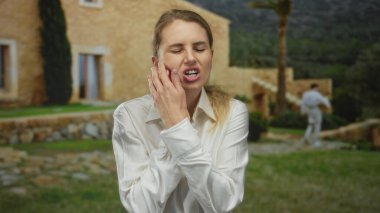 Kadın dışarıda acı çekiyor, yanağına dokunuyor, beyaz gömlek giyiyor, ağaçlar ve çimlerle dolu taş binanın yanında duruyor, kırsal alanda rahatsızlık gösteriyor..