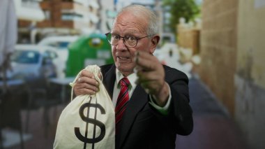Takım elbiseli kıdemli bir adam açık hava restoranındaki terasta bir dolar işaretli bir çanta tutuyor. Kalabalık bir caddede para ve yolsuzluk temaları öneriyor..