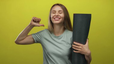 Kadın gülüyor ve yoga minderiyle kendini işaret ediyor izole edilmiş sarı arka plana karşı neşeli bir gurur ve zindelik sergiliyor.