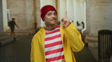 Sarı yağmurluk ve kırmızı bereli genç adam şehir sokaklarında neşeli bir şekilde el kol hareketi yapıyor. Açık hava kentsel ortamının ortasındaki şehir canlılığını ve gençlik enerjisini somutlaştırıyor..