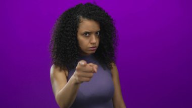 Genç İspanyol kadın, canlı mor bir stüdyo duvarına karşı çatık kaşlarıyla kamerayı işaret ediyor. Öfke..