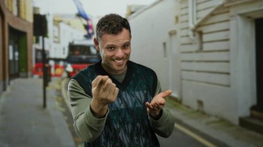 Sakallı genç bir adam, kenar mahallede kendine güvenerek gülümsüyor. Açık havada moda anlayışı sergiliyor. Olumlu hareketler yapıyor..