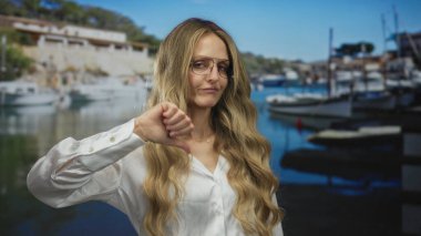 Deniz kenarında teknelerle gezen bir kadın, baş parmakları aşağıda, açık bir gökyüzünün altında manzaralı bir limanda, hoşnutsuzluğu ve yansımayı somutlaştıran bir hoşnutsuzluğu ifade ediyor..
