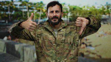 Sakallı, kamuflaj üniformalı bir asker yeşil bir parkta arka planda ağaçlarla dikilirken ve dışarıda karışık duygular gösterirken baş parmağıyla işaret veriyor..