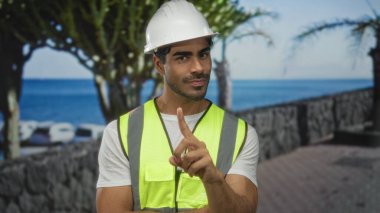 Beyaz şapka ve yansıtıcı yelek giymiş genç İspanyol mühendis parmak kaldırmayı işaret ediyor..