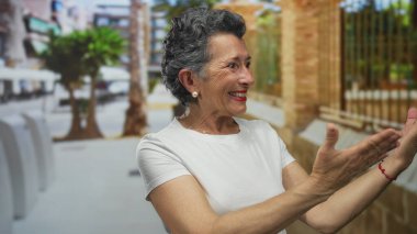 Yaşlı bir kadın, şehir sokaklarında elleri ile mutlu bir şekilde el kol hareketi yapıyor, canlı bir kasaba manzarasının neşeli ruhunu yakalıyor..