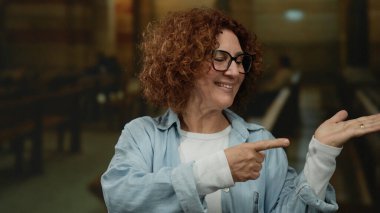 Gülümseyen ve el kol hareketi yapan bir kadın kilisenin arka bahçesinde günlük kıyafetlerle, kapalı bir dini ortamda güven ve pozitiflik gösteren bir tabela..