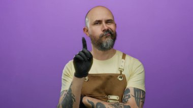 Önlüklü ve siyah eldivenli yakışıklı sakallı adam mor bir arka plana karşı duruyor, düşünceli bir ifade sergiliyor..