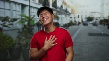 Şehrin sokaklarında neşeli bir şekilde gülen genç bir adam kırmızı tişört giyip açık havada mutluluk ve enerjiyi somutlaştırıyor.