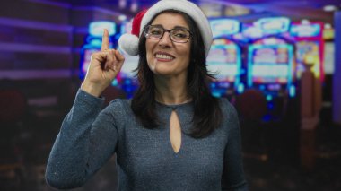Kumarhanede kırmızı Noel Baba şapkası takan kadın gülümseyerek slot makinesini işaret ediyor..