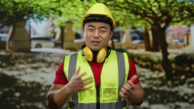 Emniyet yeleği ve kasket takan Asyalı genç adam ağaçlarla ve şehir manzarasıyla güvenle gülümsüyor..