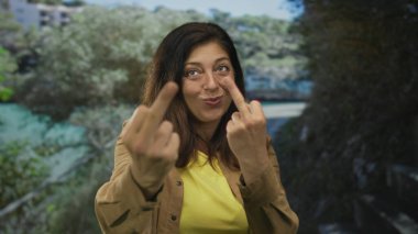 Ormanda, yeşil ağaçların arasında, kameraya göz kırparken orta parmağını sallayan kadın, hem orta parmağını kaldırmış hem de odaklanmış;.