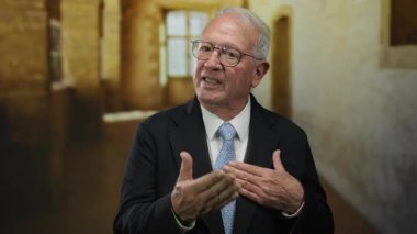 Takım elbiseli kıdemli bir adam eski bir üniversitede duruyor, elleri yüreğinde, akademik bir ortamda bilgelik ve duygu yayıyor, iş ve miras karışımı bir şey yaratıyor..