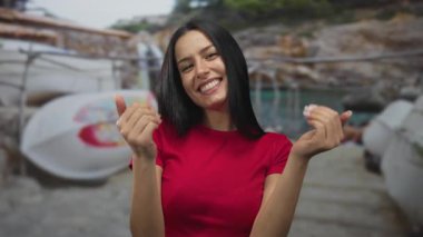 Kırmızı jestli genç İspanyol bir kadın arka planda kano ve deniz manzaralı bir plaja gelir ve açık deniz havası yansıtır..