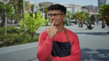 Kırmızı kazaklı ve gözlüklü genç bir adam şehir sokaklarında dikiliyor. Başparmağını kente odaklanmış ifadesiyle işaret ediyor..