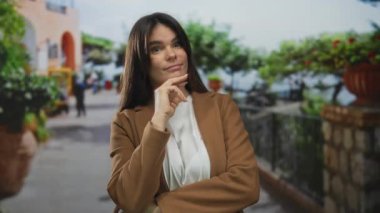 Canlı bir şehir caddesinde duran genç bir kadın kahverengi ceketli düşünceli bir ifade ile arka plan yeşillikleri sergiliyor..