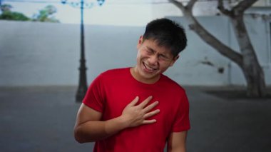 Şehir caddesinde gülümseyen genç adam açık şehir ortamında ağaçlar ve lamba direğiyle kırmızı tişört giyiyor..