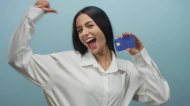 İspanyol kadın gülümseyip kendini işaret ederken mavi bir kredi kartını mavi bir duvarın arkasında tutarak kendine güvenini ve neşesini ifade ediyor..