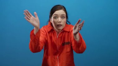 Turuncu hapishane tulumlu esmer kadın, mavi bir arka plana karşı anlamlı jestler yapıyor, karmaşa ve tefekkür duygularını somutlaştırıyor..