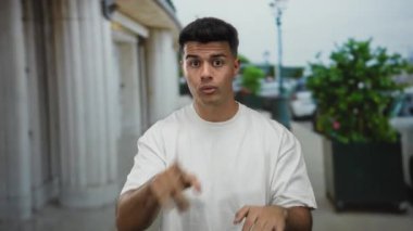 Beyaz gömlekli genç adam hareketli şehir caddesinde köşe yazıları ve yeşilliklerle dolu, şehir ortamında ifade ve etkileşim sergiliyor..