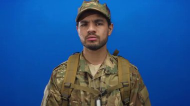 Kamuflaj üniforması giyen İspanyol adam, mavi stüdyoda sıkı sıkıya kollarını kavuşturuyor..