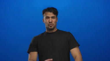 Siyah tişörtlü, korku dolu yüzlü genç bir adam izole edilmiş mavi arka planda duruyor, minimalist bir alanda korku ve ihtiyatı yakalıyor..