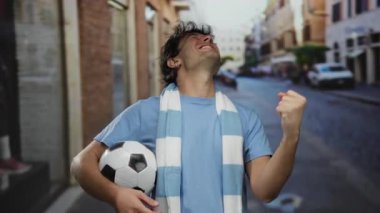 İspanyol bir adam elinde futbol topuyla sokakta kutlama yapıyor, mavi gömlek ve beyaz bir eşarp giyiyor, spor zaferi için sevinç ve heyecan ifade ediyor..