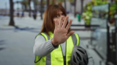 Neşeli bir şehir kaldırımında neon yelek giymeyi, elinde bisiklet kaskı, bulanık palmiye ağaçları ve kafeleri olan güneşli bir arka planda.