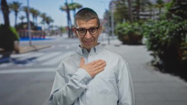 Avuç içi çizgili bir sokakta gülümseyen gözlüklü bir adam çizgili bir gömlek giyiyor ve göğsünde canlı yeşillik ve şehir manzarasıyla mutlu görünüyor..