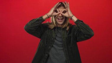 Kadife ceket giyen adam kırmızı stüdyoda dürbün yaparken gülümsüyor; merak..