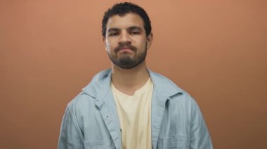 Genç İspanyol bir adam stüdyoda mavi gömlek giyen turuncu bir duvarın önünde sert bir somurtmayla kollarını kavuşturur;.